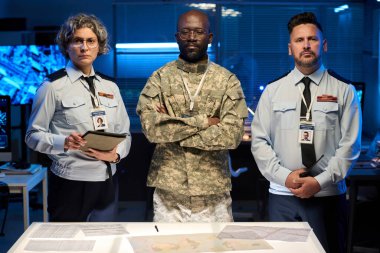 Mavi üniformalı iki yetişkin subay ve onların genç Afrikalı Amerikalı erkek meslektaşları askeri komuta ofisinde kameraya bakıyor.