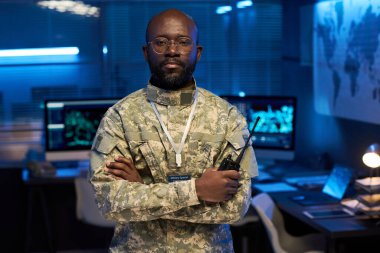 Gözlüklü ve kamuflaj üniformalı ciddi bir Afro-Amerikan subayı. Kameraya bakarken elinde telsiz var.