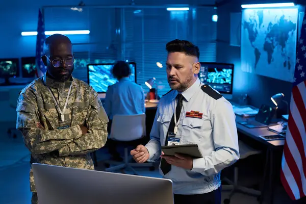 Üniformalı genç ve olgun erkek ordu subayları ortak çalışma alanındaki bilgisayar ekranında verileri tartışıyor.