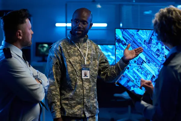 Kamuflaj üniformalı genç, kendinden emin bir subay olgun meslektaşlarına interaktif ekrandaki grafik verileri açıklıyor.
