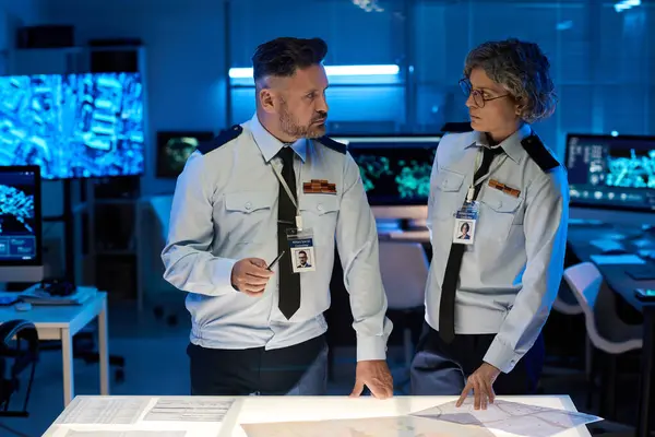 Üniformalı ve kravatlı iki kendinden emin yetişkin subay askeri komuta merkezinde evrakları tartışırken birbirlerine bakıyorlar.