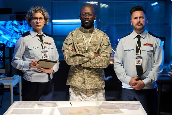 Mavi üniformalı iki yetişkin subay ve onların genç Afrikalı Amerikalı erkek meslektaşları askeri komuta ofisinde kameraya bakıyor.