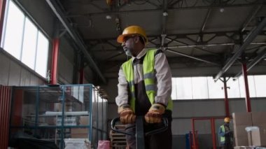 Yetişkin Asyalı teknisyen üniformalı, sarı yelek, hangarda yürürken palet vagonunu çekerken, malzeme veya hammadde taşırken orta boy bir fotoğraf.