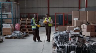 Geniş etnik çeşitlilikte erkek depo çalışanları, Asyalı yönetici ve Afrikalı Amerikan mühendis koruyucu tulumlar giyiyor, başlık takıyor, sarı yelekler giyiyor, geniş bir hangarda konuşuyor, çalışma planlarını tartışıyor.