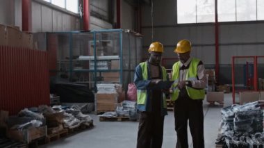 Orta boy Afro-Amerikan ve Hint erkek depo teknisyenleri, yüksek çözünürlüklü yelekleri, tulumları, panodaki şemaya bakan başlıkları, palet yerleştirme noktalarını ve