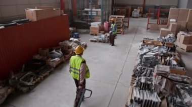 Geniş çaplı gözetleme kameraları geniş bir depoda çalışan çok ırklı erkek çalışanları gösteriyor. Kutuları kamyonla taşıyorlar, paletlerdeki malları ve bileşenleri kontrol ediyorlar.