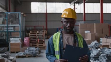 Afro-Amerikalı neşeli erkek depo elemanının orta boy portresi pano ile birlikte, tulum, kot gömlek, sarı yelek ve şapka giyiyor, depoda duruyor, kameraya bakıyor, gülümsüyor.