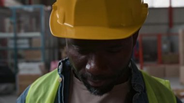 Genç Afro-Amerikan erkek depo uzmanının yakın plan portresi sarı yelek, başlığı yavaşça kaldırıyor, kameraya bakıyor, güvenle gülümsüyor, gündüz vardiyasında depoda dikilirken