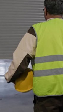 Dikey orta boy erkek depo çalışanı üniformalı, sarı görünürlük yeleği sabah işe geliyor, paten kapısını kaldırıyor, depolara girerken kaskı takıyor.