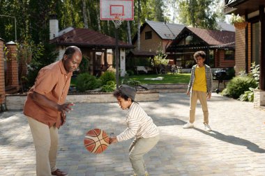 Grandfather enjoying outdoor basketball game with grandchildren in backyard of suburban home surrounded by greenery and small houses clipart