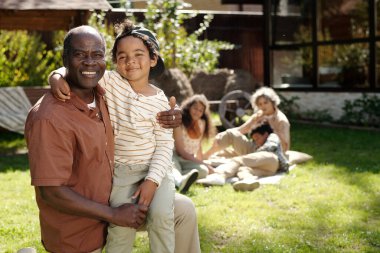 Büyükbaba aile toplantısı sırasında arka bahçede gülümserken küçük torununu tutuyor. Aile üyeleri arka planda dinleniyor, güneşli günün tadını çıkarıyorlar.