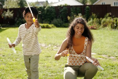 Young boy playing outdoors with his mother in lush garden on bright sunny day, both enjoying activities with bubble wands while smiling happily in natural setting clipart