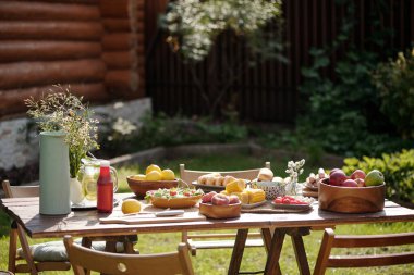 Bahçedeki ahşap masa çeşitli taze meyve ve çiçeklerle açık havada toplanmak için cazip bir ortam yaratıyor. Parlak güneş ışığı, elma, limon ve armut kaselerini vurguluyor.