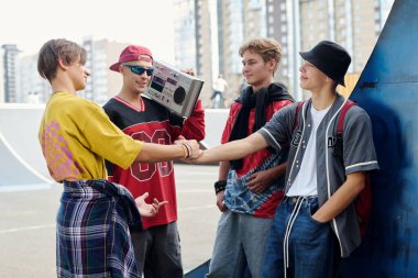 Bir grup genç kaykay parkının yanında sıradan kıyafetlerle dikiliyorlar, biri teyp kutusunu tutarken diğer ikisi el sıkışıyor. Arka planda kentsel ortam