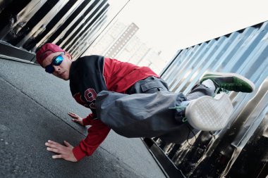 Şehir planına karşı dinamik break dans hareketi yapan şehir dansçısı, bir elinde denge kurarken kentsel kıyafetler ve güneş gözlüğü takıyor ve havada bacakları tekmeliyor.