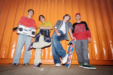 Turuncu bir duvarın önünde duran dört genç adam kaykay ve boombox ile canlı şehir kültürünü somutlaştırıyor. Gençlik enerjisi ve tarzı yayıyorlar.