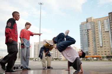 Bir grup break dansçı arka planda yüksek binalarla şehir bölgesinde etkileyici hareketler sergiliyor. Seyirciler dansçıları izleyip işaret ediyor.