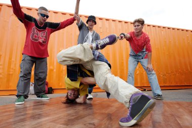 Bir grup genç adam açık havada break dans yapıyor, arka planda parlak turuncu konteynırla dinamik dans hareketleri sergiliyorlar.