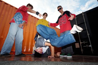 Bir grup genç, enerji ve gençlik tasvir eden renkli nakliye konteynırlarına karşı şehir sokaklarında break dans hareketleri yapıyor.