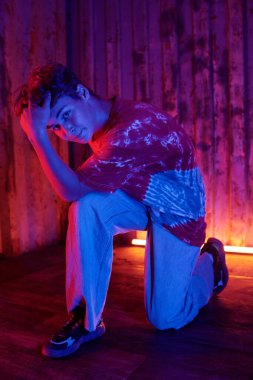 Yaratıcı iç mekanda poz veren genç bir adam karışık kırmızı ve mavi ışıklandırmalı, elleri başının üstünde ve bir dizi bükülmüş. Ahşap duvar arkaplanı fotoğrafa eşsiz bir atmosfer ekliyor