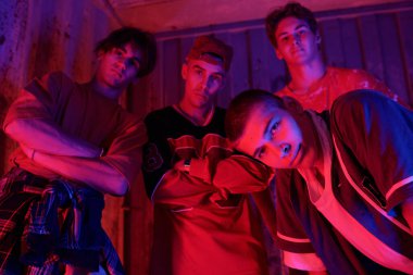 Ciddi ifadelerle poz veren ve kırsal ahşap duvarın önünde kollarını kavuşturan bir grup genç adam, loş mavi ve kırmızı ışık dramatik bir hava yaratıyor.