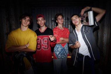 Dört genç adam, farklı kıyafetlerle gelişigüzel giyinmiş bir müzik kutusuyla birlikte duruyorlar. Kapalı bir ortamda güven içinde poz veriyorlar cesur, kentsel bir havayla.