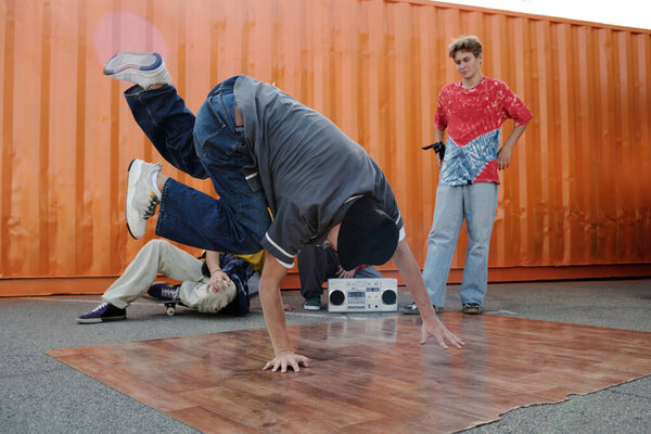 Outdoor breakdance performance with two friends watching closely. One person executing a dynamic dance move beside an orange container