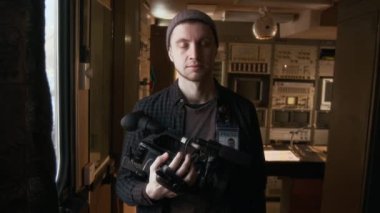 Profesyonel dijital kamera taşıyan, TV yapım minibüsünde duran, güvenle gülümseyen, günlük bere ve gömlek giyen, neşeli, yakışıklı beyaz erkek videografın orta boy portresi.