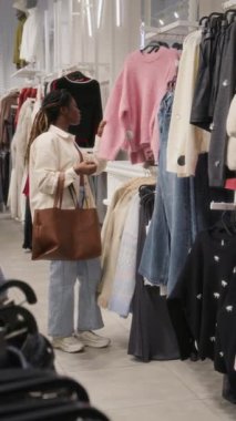 Dikey olarak, tek başına alışveriş yapan, elbise mağazasında giysili eşyalara bakan Afro-Amerikan genç bayanın fotoğrafı.