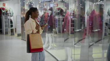 Siyahi kadın alışveriş koliklerinin kağıt torbalar ve akıllı telefonlar taşıdığı dükkanın camına bakarken ve yeni kıyafet koleksiyonunu değerlendirirken, hafta sonunu alışveriş merkezinde geçirirken.