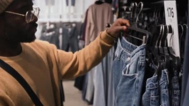 Şapkalı, gözlüklü, şık Afro-Amerikan bir adamın elbise mağazasından kot pantolon seçerken göğsünden çekilen fotoğrafı.