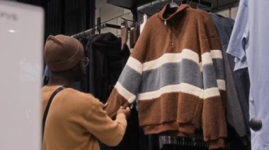 Siyah erkek müşterinin bereli ve sweatshirt 'lü arka görüntüsü. Giysi dükkanında beyaz ve gri yatay çizgili sıcak kahverengi kazak seçiyor. Malzeme kalitesini kontrol ediyor.