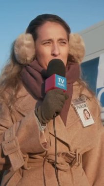 Beyaz kadın gazetecinin kulaklıklı, kışlık kürklü, mikrofona konuşan ve yerel TV kanalına haber sunan orta boy dikey görüntüsü.