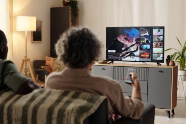 Modern oturma odasındaki bir kanepede oturmuş televizyon ekranında film seyreden iki kişi. Bitkilerle süslenmiş ve şık mobilyalarla süslenmiş bir yerde.