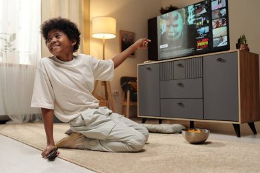 Afrika kökenli Amerikalı bir adam, elinde uzaktan kumandayla gülümserken arka planda televizyon ve mobilyalarla yerde oturuyor.