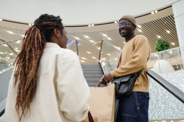 Giyim kuşamında mutlu bir Afrikalı genç adam, Ticaret Merkezi 'nde yürüyen merdivende sevgilisine bakıyor.
