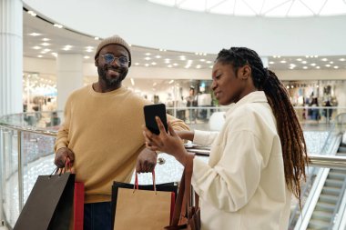 Günlük kıyafetli genç bir kadın alışveriş merkezini ziyaret ederken sevgilisini ya da kocasını internet üzerinden akıllı telefon ekranında gösteriyor.