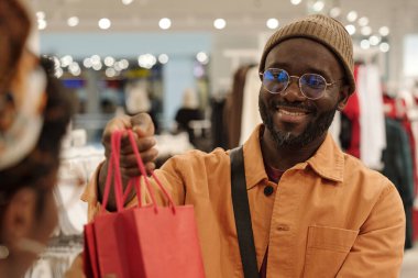 Gözlüklü ve bereli genç bir müşteri yeni kıyafetler alırken tezgahtara kırmızı alışveriş çantası uzatıyor.
