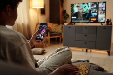 Televizyon izlerken ve telefon bildirimlerini kontrol ederken evde dinlenen biri. Yumuşak ışıklandırmalı ve kucağında patlamış mısırla rahat bir atmosfer.