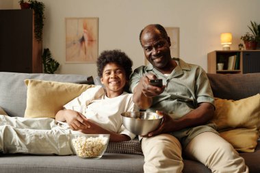 Çocuk ve yaşlı adam kanepede oturuyor, uzaktan kumandalı ve patlamış mısır tutuyor, gülümsüyor ve kaliteli zaman geçiriyor. Dekoratif duvar sanatı ve sıcak ambiyansı olan rahat bir oturma odasında dinleniyor.