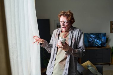 Elinde kahve olan, oturma odasında durup pencereden dışarı bakan garip, genç bir erkek zombi resmi.