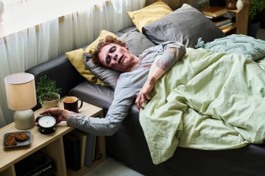 Yüksek açılı genç ölümsüz erkek çift kişilik yatakta uzanıyor çalar saate uzanıyor, sabah uyanıyor.