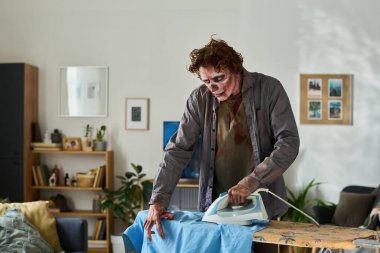 Genç zombi erkeğin sabah mavi gömleğini ütülerken orta boy bir pozu.