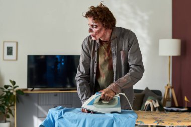 Sabahları oturma odasında mavi gömleğini ütülerken elini yakan cahil erkek zombi resmi.