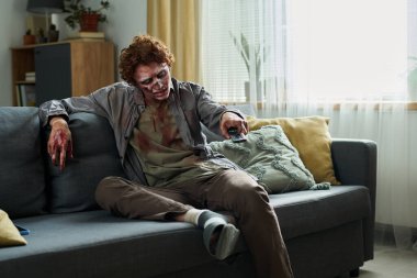 Young male zombie sitting relaxed on couch using remote control to choose TV show clipart