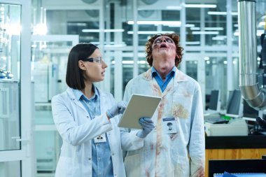 Medium shot of young Caucasian female scientist working with tablet in laboratory, speaking to undead male colleague, noticing him standing in stupor clipart