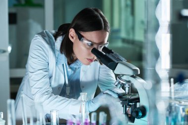 Genç beyaz kadın bilim adamı gece geç saatlerde biyokimya laboratuvarında reaktörlerle çalışırken modern mikroskop kullanıyor.