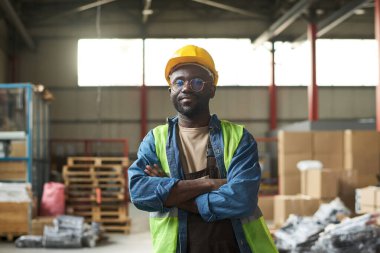 Kasketli, tulumlu, kot gömlekli ve floresan ceketli genç bir fabrika işçisi geniş bir depoda kameraya bakıyor.