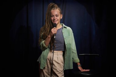 Uzun saçlı, elinde mikrofonla sahnede duran gülümseyen bir kadının portresi. Sahne performansı sırasında kendinden emin ve rahat görünüyor