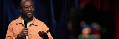 African American man wearing glasses holding microphone while delivering speech on stage bathed in soft light. Background features dark colored curtains enhancing focus on speaker clipart